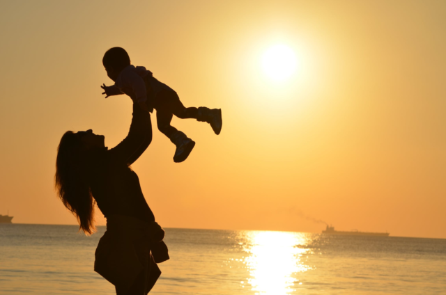 A sunset over the ocean casts a silhouette around a mother lifting her child above her head.