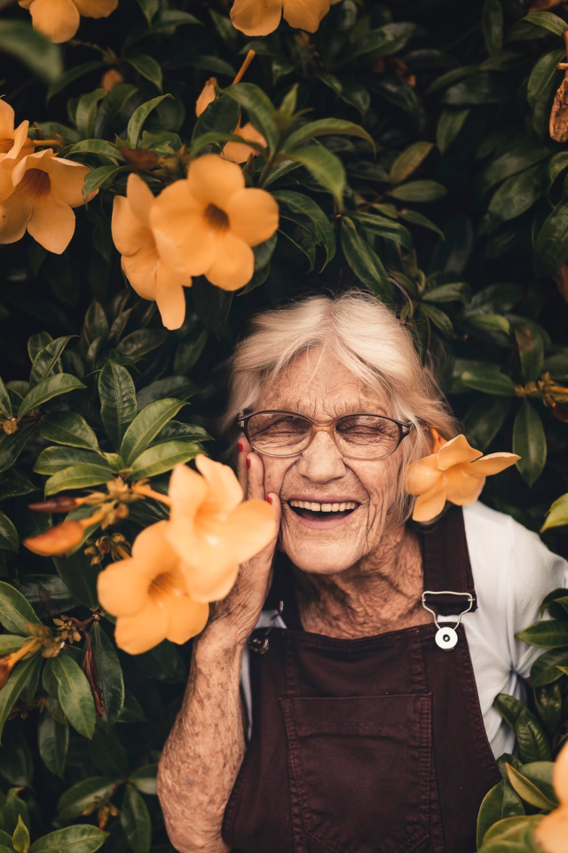 A grey-haired woman smiling widely, standing amid vines with yellow flowers.