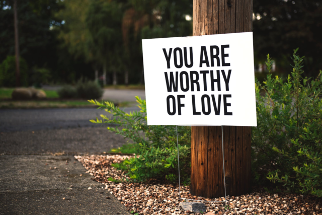 A white sign with black text reading “You are worthy of love” stands next to a wooden pole off a forest trail.