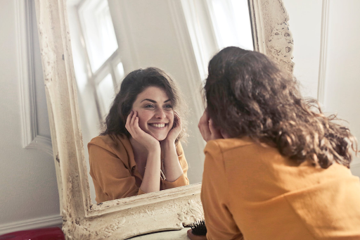 A woman wearing a yellow shirt looks at her reflection in a mirror, smiling