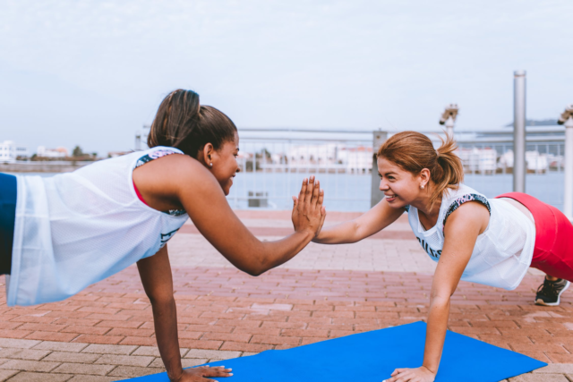 Two women in athletic apparel face each other and share a high-five while holding a plank position.
